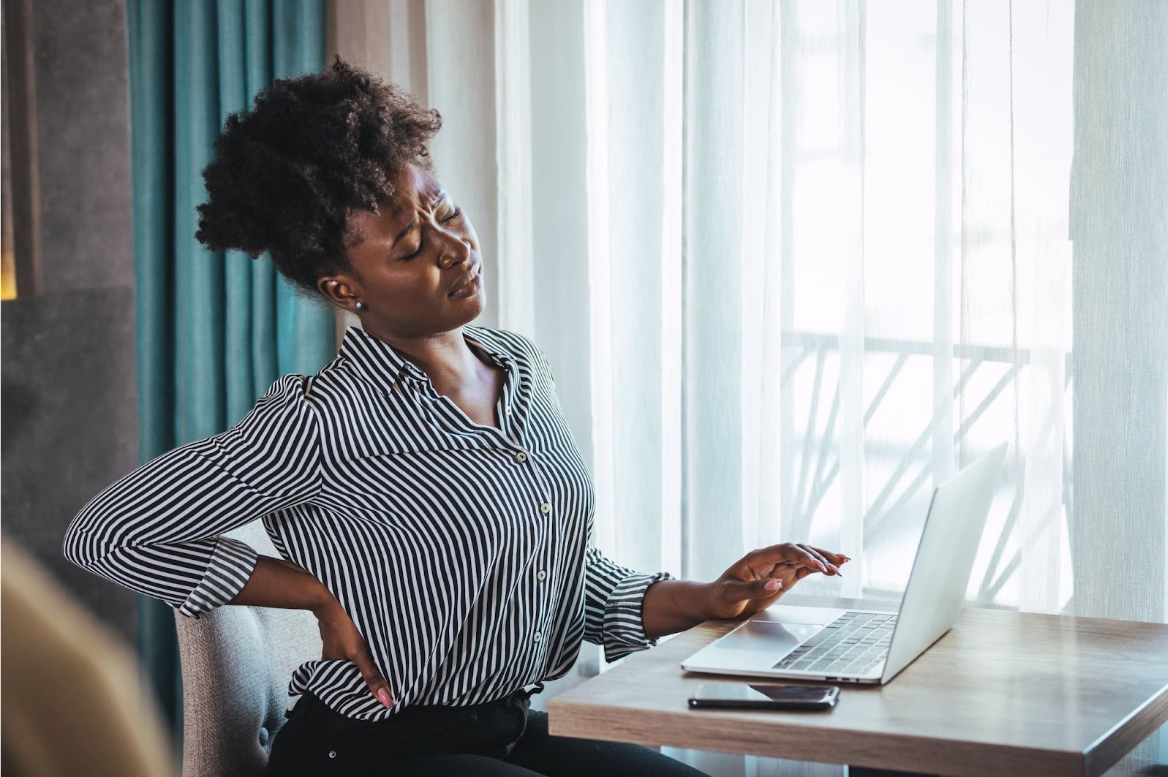 A woman sits at her desk in front of her laptop, holding her painful lower back.