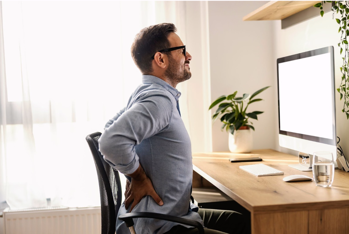 Man sitting at a desk, leaning back in his chair and holding his lower back in discomfort.