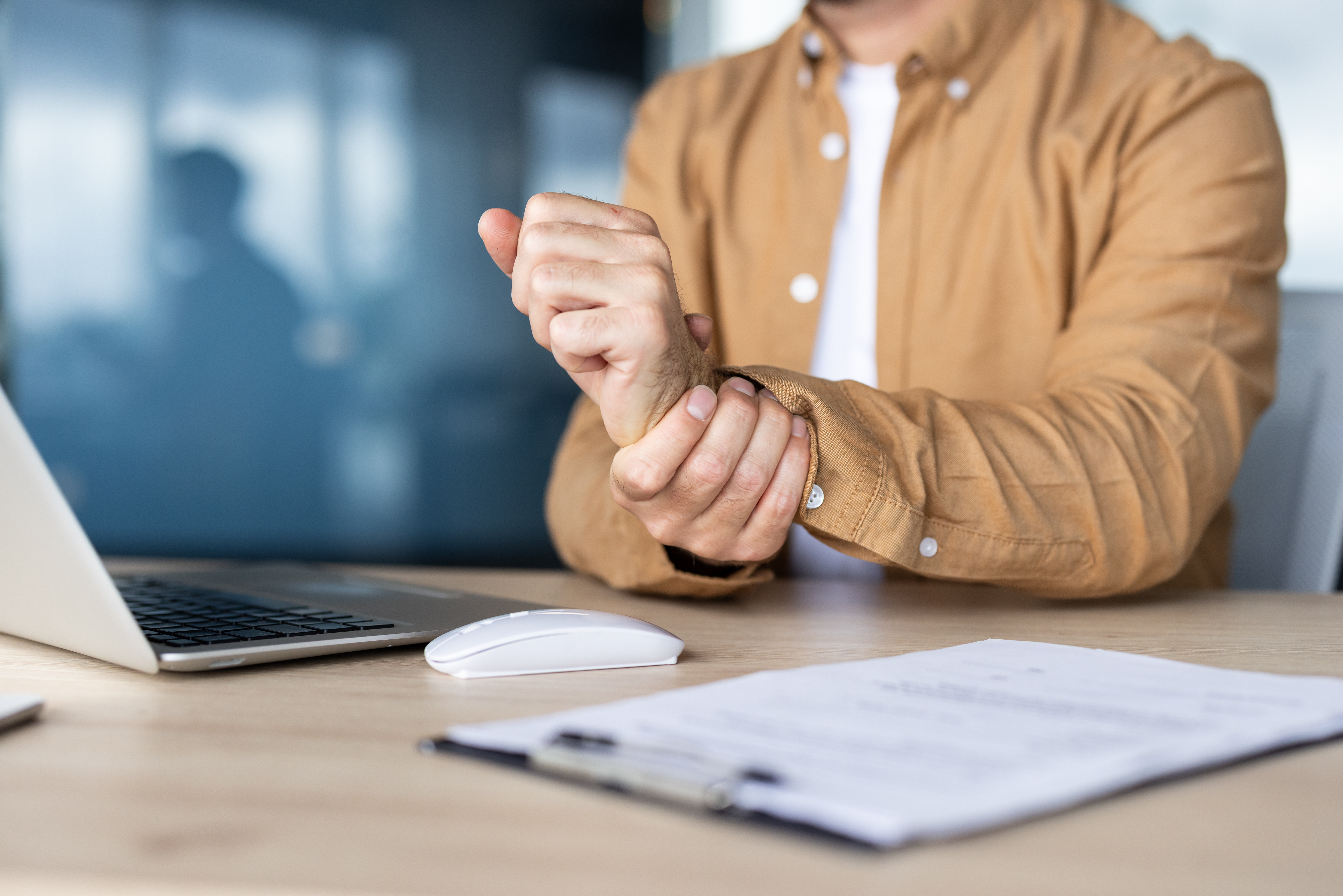 Businessman suffering from wrist pain while working on laptop at desk in office, possible carpal tunnel syndrome or other repetitive strain injury caused by prolonged computer use