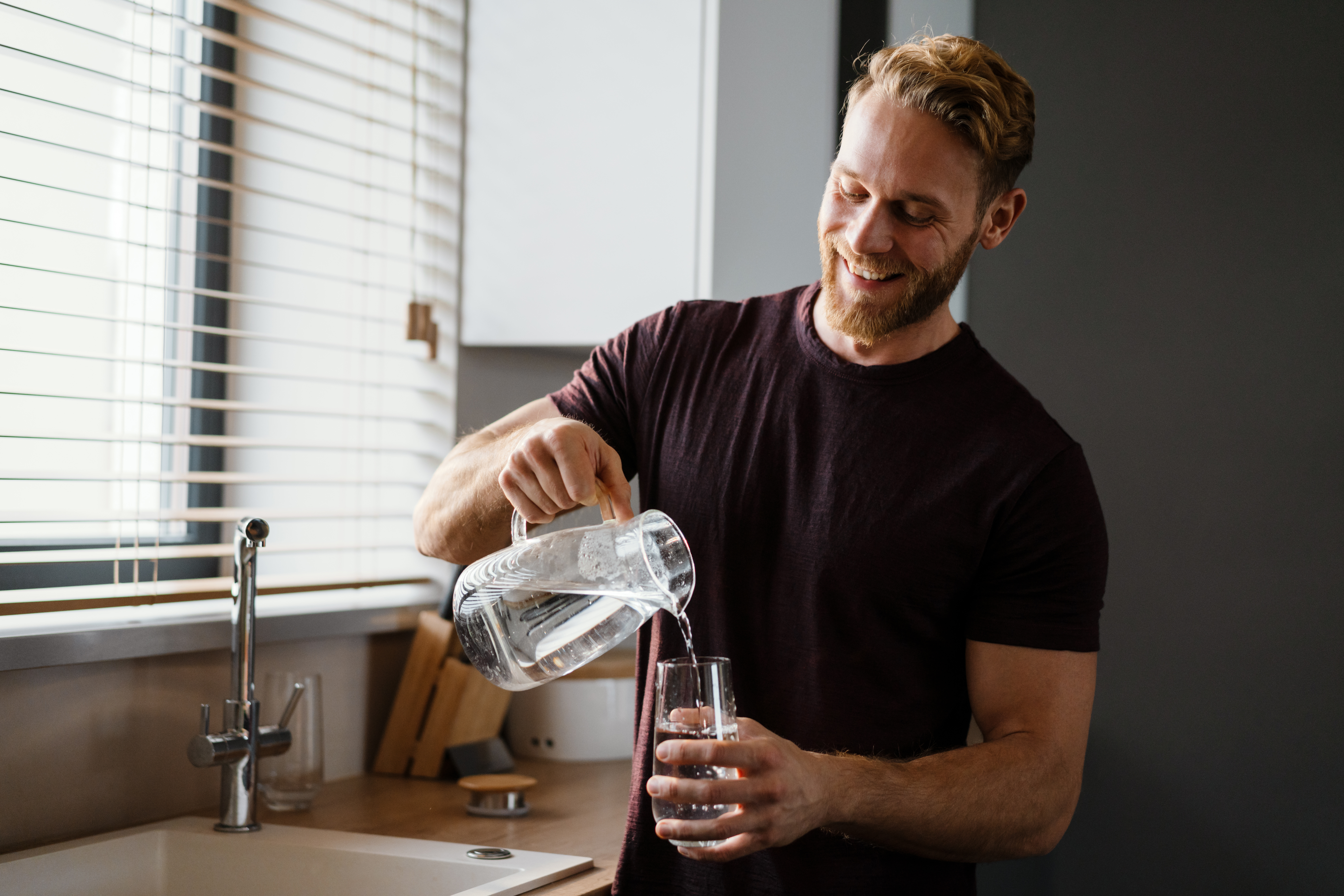 Attractive young man pouring water from a juf in a glass while standing at the kitchen sink