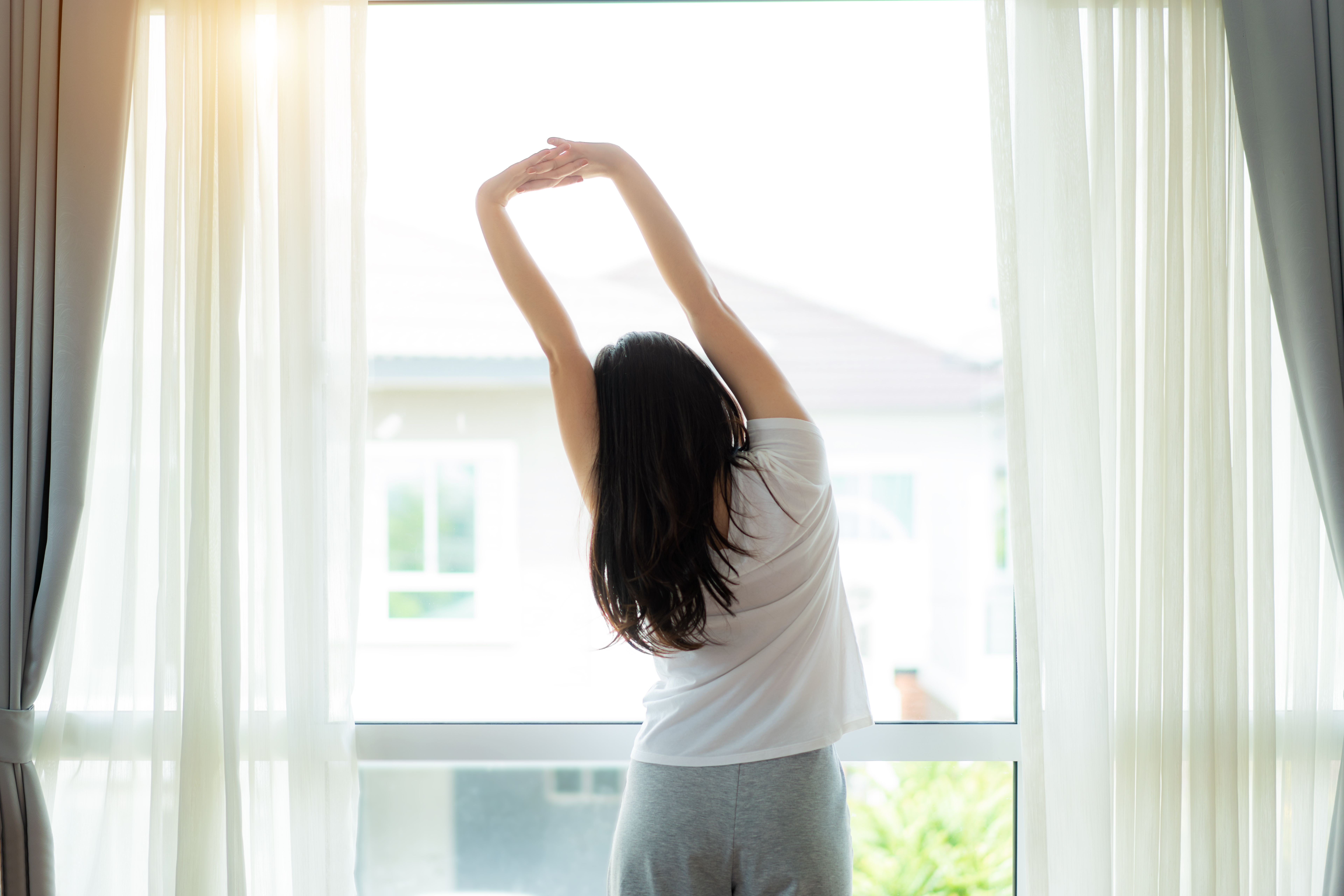 Rear view of Asian Woman stretching hands and body near window after wake up in bedroom at home. Concept for start new day with happiness. Young happy working female life